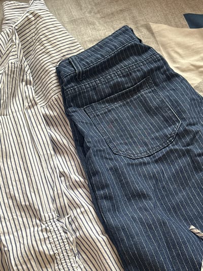 The image shows a close-up of clothing items on a bed. A pair of dark blue, striped shorts with a visible pocket are on top of a white and blue striped shirt. The background consists of a patterned bedspread. There is no indication of the city of Agarak, Armenia, or any other location information in the image.