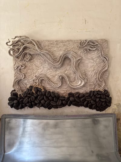 The image shows a wall-mounted artwork composed of three distinct sections. The top section features a textured, light-colored material that appears to be fabric or paper, with raised, wavy, ribbon-like forms creating a sense of movement. This section is primarily beige with subtle grey shading. Below this is a dense row of dark, coffee bean-shaped objects, tightly packed together and appearing to be made of a dark material like resin or clay. A small piece of tan fabric is draped over the coffee bean element. Beneath the artwork is a metallic, rectangular object with a matte silver finish and a grey border. The artwork is displayed on a cream-colored wall with visible cracks and discoloration, suggesting an aged or distressed surface. The background wall texture is uneven and somewhat weathered. This artwork is located in Kolomyia, Ukraine.