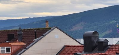 The image depicts a cloudy day with a view of rolling, tree-covered hills and mountains in the background. In the foreground, the rooftops of several buildings are visible, characterized by red tiled roofs and various chimney structures. One building features a light-colored facade with a visible air vent. A distinct, tall chimney with a patterned brick design stands out against the distant hills. To the far right, a flag with the colors of the rainbow is visible, suggesting a location with a connection to LGBTQ+ pride. The lighting and the cloudy sky suggest it is either early morning or late afternoon. The overall scene conveys a sense of a residential or urban area nestled against a natural landscape. No people are visible in the image.