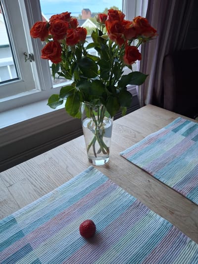An indoor scene depicts a bouquet of orange roses in a clear glass vase filled with water, positioned on a light-colored wooden table. Two placemats with thin, multicolored stripes in shades of blue, green, pink, and yellow are laid out on the table. One strawberry rests on the lower placemat. In the background, a window reveals a view of buildings and the sky. The image is shot from a slightly elevated angle. The context provided places the scene in Åsgårdstrand, Norway.