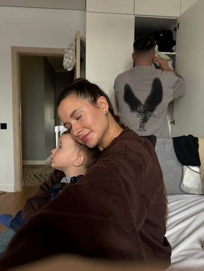 The image captures an intimate indoor family scene, rotated 90 degrees clockwise from its natural upright orientation. In the foreground, a woman with dark hair is seen from the chest up, her head gently resting against a young child's head. Her eyes appear closed or cast downward, and she has a subtle, content smile, suggesting a moment of tenderness or affection. She wears a dark brown long-sleeved top and has a small silver hoop earring in her left ear. The child, likely a toddler, is nestled close to the woman, looking to the side with a calm, slightly pensive expression. The child is wearing a dark top with some grey and black straps or harness visible.

In the mid-ground, a man is visible, leaning into an open white cabinet or closet. He is seen from the back and side, wearing a light grey long-sleeved shirt with a dark graphic design on the back that depicts what appears to be a bee or insect with wings, accompanied by the text "BE POSITIVE". He seems to be arranging or retrieving items from the cabinet, which contains various folded clothes.

The background shows white cabinetry that extends upwards and to the left. To the right, a bed or sofa is partially visible, covered with white bedding, including a textured white blanket or sheet. The flooring appears to be light-colored wood. The overall setting suggests a casual home environment, possibly a bedroom or living area. The lighting is soft and even, indicating an indoor setting, likely during the day or under artificial light. The image conveys a warm, personal moment shared between family members.