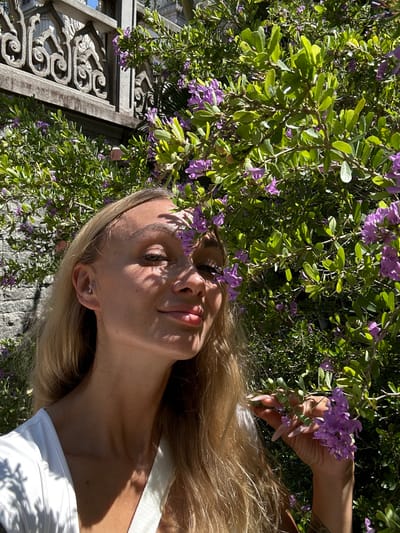 A woman is lying down under a flowering bush. The bush has green leaves and purple flowers. The woman has long blond hair. She is wearing a white shirt. There is a carved stone structure in the background. The location is likely outdoors, possibly in a garden or park in Dĩ An, Vietnam.