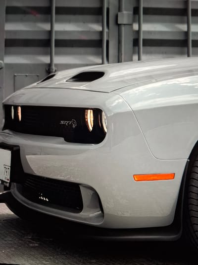 A white Dodge Challenger SRT Hellcat sits in what appears to be a garage or industrial setting, indicated by the corrugated metal wall in the background. The car's front is prominently featured, with its distinctive black grille, red SRT badge, and illuminated headlights. The hood has two air vents. A license plate with "797 RUSSIA" is visible on the left. The car is parked on a textured surface, possibly a ramp or a metal grate. An orange reflector is on the car's front fender. The overall lighting is dim, with highlights on the car's paint and a spotlight effect on the headlights. There are no people visible in the image. The scene is devoid of any context that would place it specifically in the city of Argun, Russia, other than the license plate inscription.
