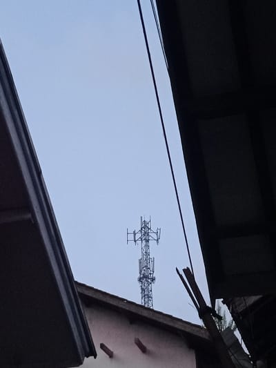 The image depicts a static scene in Ciputri, Indonesia, observed from a low angle looking upwards. Dominating the upper two-thirds of the frame is an overcast, light blue-grey sky. Centrally positioned against this sky is a tall, metal lattice telecommunications tower equipped with multiple antenna arrays and communication platforms.
The foreground is framed by dark, angled architectural structures. On the left, a dark grey underside of an eave or roofline extends from the upper left corner diagonally downwards. On the right, a dark, overhanging structure is visible, with a thin black utility line traversing diagonally from top right towards the center. Below this utility line, partially obscured by the dark structure, is a fractured, dark-colored object, consistent with broken wooden debris or a similar structural fragment.
Below the cell tower and extending across the lower portion of the image is the upper section of a building. Its light-colored (off-white or light pink) facade features two small, horizontally rectangular, dark brown protrusions. Above this facade, a dark-tiled or similarly colored roofline is visible, connecting to the structures that frame the image. No people are visible. No actions or interactions are occurring.