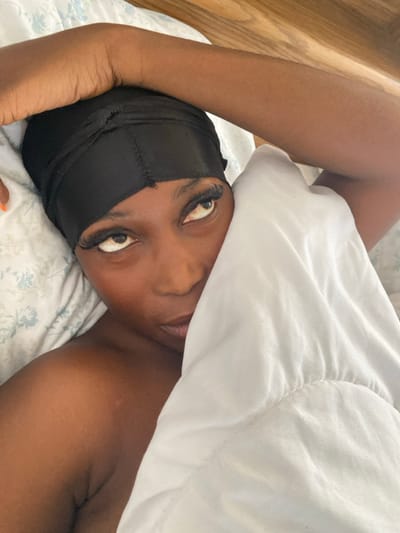 A single individual is depicted in an indoor setting, likely a bedroom. The individual, appearing to be an adult female with dark skin, is lying down and looking upwards with her eyes rolled. She wears a black satin head wrap. Her left arm is bent at the elbow, with the hand positioned over her head. A white quilted comforter partially covers her face, exposing her left eye, nose, and mouth, as well as her right shoulder and upper arm. The bedding includes a pillow with a light-colored, subtly patterned pillowcase. A wooden surface, possibly a headboard or wall, is visible in the background. The specific location in Tafi, Ghana, cannot be determined from the visual information provided.