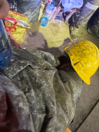 An indoor scene in Chelopech, Bulgaria, depicts a dusty industrial or mining environment. In the foreground, the lower body of an individual is visible, dressed in olive-green workwear heavily coated with a light, powdery residue. A blue plastic water bottle is partially visible on the left side of the frame. A scuffed yellow hard hat, bearing dark, indistinct markings consistent with handwriting (e.g., "KVK" or similar), rests on the right leg. In the mid-ground, the lower legs and feet of a second individual, clad in dark work boots with reflective ankle bands, are visible on a soiled floor. The immediate surroundings include a large black plastic bag, a yellow plastic bag, and another blue plastic water bottle on the ground. The floor surface exhibits varied textures, including dirt, concrete, and a section of metal grating in the lower right. The scene is illuminated by artificial, yellowish light, highlighting the pervasive dust and debris.