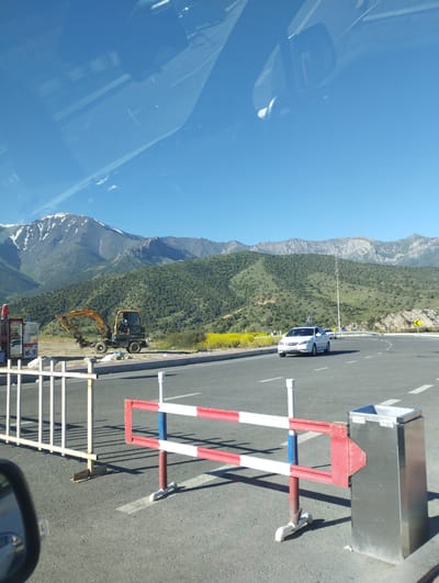 The image depicts a roadside scene with mountains in the background, suggesting a location in Dardar, Uzbekistan. In the foreground, a road is visible with a white car driving on it. A red and white striped barrier is placed on the side of the road, with a silver bin next to it. To the left of the road, a yellow excavator is parked on a dirt patch. The mountains are covered in green vegetation, with some snow-capped peaks visible at the top. The sky is clear and blue, indicating a sunny day. The overall impression is one of a scenic, rural area with some ongoing construction work.