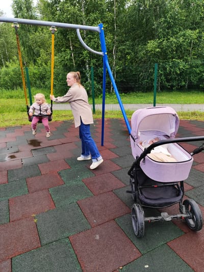 A woman is pushing a young child on a swing at a playground. The child is wearing a light-colored jacket and pink pants, while the woman is wearing a beige jacket and blue jeans. Next to the swing set is a pink baby stroller with a baby inside. The playground has a rubberized surface made of red and green interlocking tiles. In the background, there are trees and a green metal fence. The sky is overcast, suggesting a cloudy day, and there are puddles on the ground, indicating recent rain. The overall setting appears to be a park or outdoor recreational area. The woman seems to be enjoying her time with the child on the swing, and the scene depicts a family outing. There is no visible text in the image.