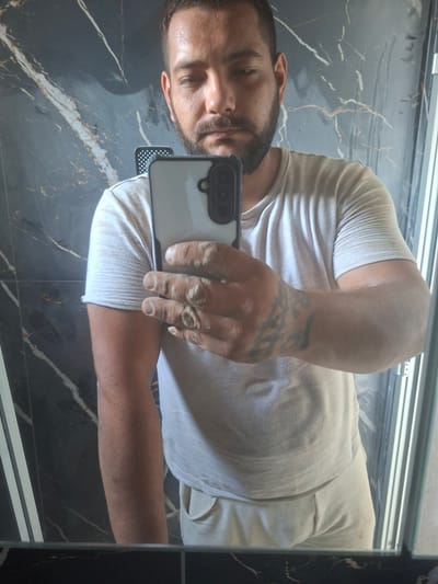 The image shows a man taking a mirror selfie. The man is wearing a light-colored t-shirt and appears to have a beard. He is holding a smartphone in his right hand, angled to capture his reflection. His left arm is visible, and he has a tattoo on his forearm. The background is a dark, marble-like surface, possibly a countertop or wall. A thin, horizontal metallic element is visible at the top of the frame. The lighting suggests it could be daytime or well-lit indoor environment. The image is taken in Dolna Malina, Bulgaria.