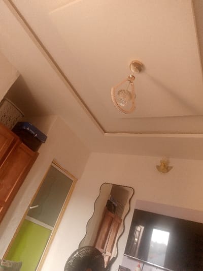 A low-angle shot captures an indoor space. A tan ceiling has a recessed center, a modern chandelier, and a wall fixture. Below, a wardrobe sits beside a window with green and clear glass. A blue box rests on top. A wavy-edged mirror reflects the wardrobe. A black fan base stands in front of a TV. The walls are a light tan color.
