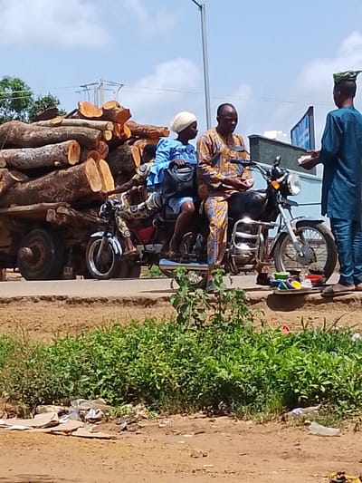 The image depicts a scene in Oke Iho, Nigeria, on a bright, sunny day with a partly cloudy sky. The primary focus is on a motorcycle carrying three people and a large load of freshly cut logs. A man in a patterned yellow and brown outfit is driving, with a woman in a blue outfit and headscarf seated behind him. Another person, a man in a blue outfit and a distinctive green head covering, is standing to the right of the motorcycle, holding a small stack of items, possibly goods for sale. He appears to be interacting with the man on the motorcycle.

To the left of the motorcycle, a large truck is piled high with a substantial quantity of thick, rough-hewn logs. The truck's tires and part of its chassis are visible. The motorcycle and the truck are positioned on a dirt road, with a strip of green vegetation and some scattered debris in the foreground. In the background, a building with a distinctive architectural feature and some power lines can be seen, suggesting an urban or semi-urban environment.

The overall impression is one of daily life and commerce, likely transport of harvested lumber and possibly a roadside transaction. The clear sky and bright lighting indicate it is daytime, likely late morning or early afternoon. There is no visible text in the image. The people appear calm and engaged in their respective activities.