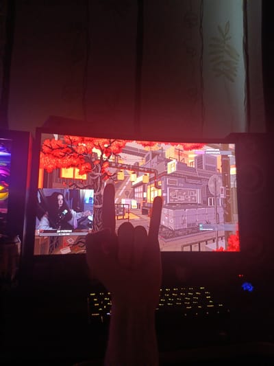 A person's hand forms a rock-and-roll gesture in the foreground, obscuring part of a computer monitor. The monitor displays a pixelated cityscape with red foliage and a smaller window showing a woman speaking into a microphone, with Cyrillic text below her. The background shows a wall with a curtain, partially illuminated by the monitor's glow. The scene is dimly lit, with some colorful LED lighting visible on the left side, likely from a computer case. The location is identified as Shipunovo, Russia.