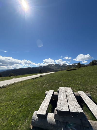An eye-level, wide-angle shot captures a scenic view from a wooden picnic table in the mountains near Cherna Mesta, Bulgaria. The sun shines brightly in the upper left corner, casting a lens flare and illuminating the clear blue sky dotted with fluffy white clouds. In the middle ground, a paved road winds through a lush green meadow with scattered yellow wildflowers, leading towards a dense forest that covers the slopes of distant mountains. The peaks of these mountains are capped with snow, creating a striking contrast against the dark green trees. The foreground is dominated by the rustic wooden picnic table, its weathered planks and log supports creating a natural and inviting seating area. The overall scene depicts a tranquil mountain landscape on a sunny day, ideal for outdoor recreation.