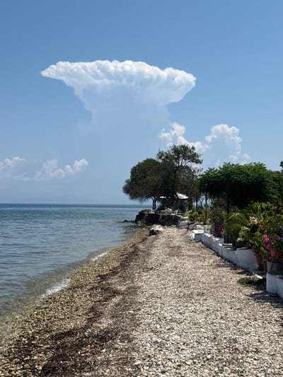 This media file shows a seaside scene with a pebble beach and the sea. A distinctive, tall, fluffy cloud with a pointed tip is visible in the clear blue sky, contrasting with smaller, scattered clouds.
The beach is covered in light-colored pebbles and seaweed. Beyond the beach, there are trees and some potted plants with pink and white flowers. White tables and chairs are set up along a white wall, suggesting a seating area for a cafe or restaurant. Streetlights are also present along the path. The sea is calm with gentle ripples.
The lighting suggests it is daytime, likely late morning or afternoon, given the shadows cast by the trees and the brightness of the sky. The weather appears to be clear and sunny. The location is identified as the Municipal Unit of Meliteieis, Greece, and the scene evokes a relaxed, summery atmosphere. There is no visible text in the image.