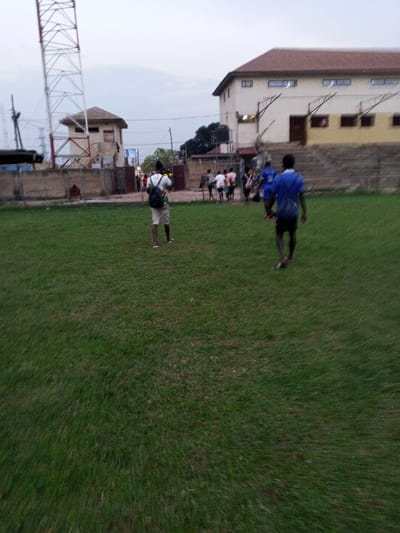 A grassy field stretches out towards a distant building. Several people are gathered near the gate. Two men, one in a blue shirt, are walking on the field. A metal tower stands tall in the background near a small building. The scene suggests an outdoor gathering on a sports field or park.