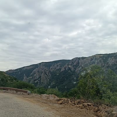 A rural road winds through a mountainous landscape under an overcast sky. The road surface is gravelly, with a rusted guardrail bordering the left side. Beyond the road, the terrain slopes upward, covered in dense green vegetation and rugged rock formations that characterize the mountains. The sky is filled with heavy, grey clouds, suggesting an impending or recent rain. The scene appears to be captured from a vehicle traveling along the road, offering a view of the natural environment of the region surrounding Madzharovo, Bulgaria.