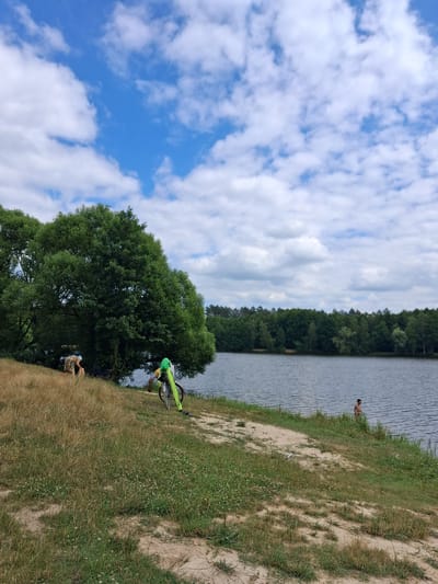 This media file depicts a sunny day at a lakeside in Reutyntsi, Ukraine. The scene is characterized by a body of water, a tree-lined shore, and a grassy, sandy bank. In the foreground, a person sits on the grass, and a bicycle with a bright green frame is propped up. Further back, on the sandy bank closer to the water, another person is visible near the shoreline. The sky is a vibrant blue, dotted with fluffy white clouds, suggesting fair weather. The presence of lush green trees and the calm water indicate a natural, possibly recreational, setting. The overall impression is one of a peaceful outdoor environment, likely during the daytime, with people enjoying the natural surroundings. There is no visible text in the media file.