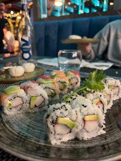 The image presents a close-up, slightly tilted view of a sushi platter served in an indoor restaurant setting. The central focus is a generous portion of various maki rolls arranged on a dark, textured plate with concentric ring patterns.

The sushi includes several types: prominent on the right are inside-out rolls (uramaki) featuring white rice speckled with black sesame seeds on the exterior. These rolls are filled with imitation crab stick and avocado. To the left, other rolls are adorned with thin slices of raw fish, including salmon, a lighter white fish, and possibly tuna, draped over the rice and fillings, which also appear to contain avocado and imitation crab. A vibrant green herb, likely cilantro or parsley, is used as a garnish.

Adjacent to the main sushi plate, on a separate, elongated dish, are two small, white, spherical food items. In the blurred background, a person is partially visible, wearing a light-colored top over a blue, padded garment, with their hand holding a golden, flat object. A clear glass sits on a reflective surface. The background further reveals blurred blue elements, possibly seating or decor, and glowing artificial lights that suggest an evening atmosphere. Reflections of lights and a hand are visible on the glossy foreground surface, where the reversed letters "7OO" can also be seen, possibly part of a logo or signage. The scene captures a dining experience in Basiglio, Italy.