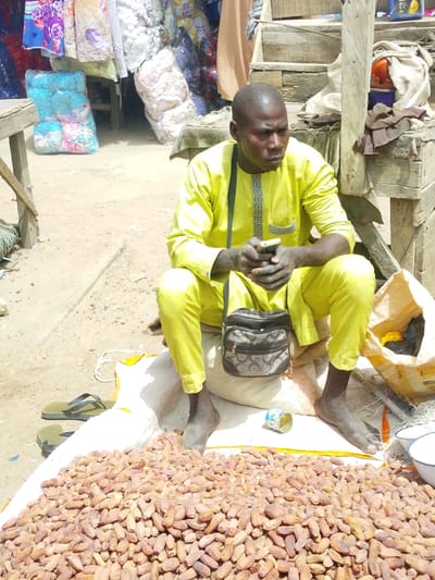 In Fagge D1, Nigeria, a sole adult male with a dark complexion and bald head is depicted seated on the ground. He wears a bright yellow tunic featuring a patterned placket and matching trousers, and is barefoot. A dark brown patterned shoulder bag is slung over his left shoulder. He holds a white-cased smartphone with both hands, looking towards the right of the frame. He is positioned on a white sack. In the foreground, a large pile of brownish-red dried dates is spread on another white sack. A crushed silver-colored can lies near his right foot, and a pair of dark-colored flip-flops are to his left. Partially visible to his right are a yellow sack and two small white bowls. The ground is exposed light brown dirt. The background reveals market activity: various bundled textiles and bedding in multiple colors are stacked and hung to the left, while a rustic wooden structure with draped fabric and an orange object is visible to the right, adjacent to a large paper sack.