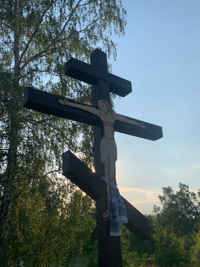 A wooden cross, depicting the crucifixion of Jesus Christ, stands outdoors under a clear sky with dappled sunlight. The cross is dark brown and features an inscription of Jesus, rendered in a light, possibly white or beige color. A white cloth with a blue embroidered pattern hangs from the lower part of the cross. The cross is positioned in front of a backdrop of lush green trees, with one prominent birch tree to the left. The setting appears to be in Mala Rublivka, Ukraine.
