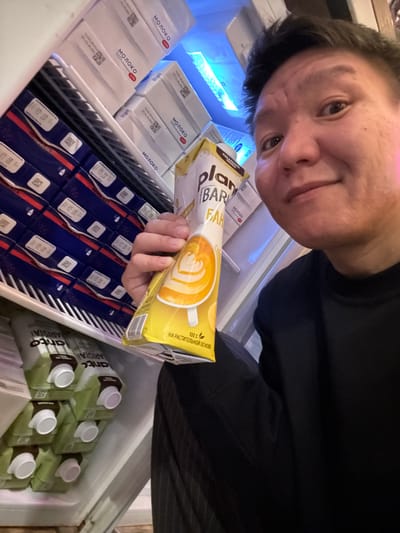 A man is taking a selfie in a refrigerator stocked with cartons of milk and plant-based drinks. The man is smiling and holding up a carton of what appears to be a plant-based coffee creamer with a picture of latte art on it. The refrigerator contains rows of white cartons labeled "МОЛОКО" (MILK) in Russian and green cartons with white caps. The image appears to have been taken inside a residential refrigerator, potentially in the city of Veshki, Russia, given the presence of Russian labeling.