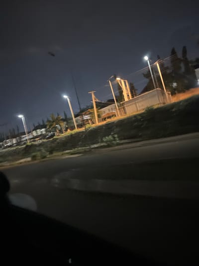 The image depicts an urban street scene at night, potentially in or around Jiru, Nigeria. A row of streetlights illuminates a road bordered by vegetation on one side and what appears to be buildings further back. Utility poles with wires are visible, suggesting an infrastructure for electricity and communication. The image is taken from the perspective of being inside a vehicle. The light is dim, typical of nighttime conditions in a city.