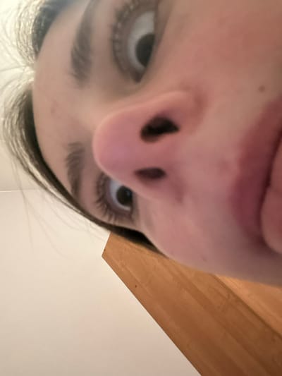The close-up shot from below reveals a person's face dominated by their nose and eyes. The nostrils are prominently displayed. Above the nose, the upper lip is visible. The eyes, framed by dark eyelashes, gaze upwards. Eyebrows are dark and thick. Some hair strands are visible near the jawline. A blurred light brown wooden surface appears in the upper right corner, contrasting with the light wall or ceiling in the lower right.