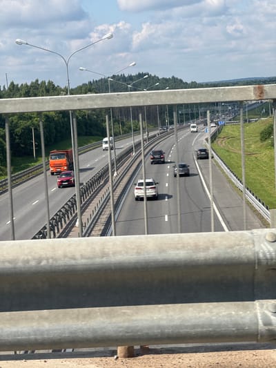 This image depicts a view from a highway overpass in Poroshkino, Russia. The scene shows a multi-lane highway with several cars and a truck driving on it. The highway is bordered by a grassy embankment on one side and a dense forest on the other. In the foreground, metal railings partially obscure the view of the highway, suggesting the photograph was taken from a bridge or walkway. Streetlights are visible along the side of the road, indicating it's a well-maintained route. The sky is partly cloudy, suggesting it's daytime. The overall impression is a typical highway scene in a semi-rural or suburban area. There is no visible text in the image.