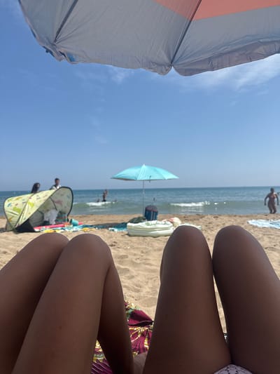 The image shows a first-person perspective of two legs resting on a beach, with a beach umbrella in the foreground and the sea and sky in the background. The beach appears to be sandy, with some towels and a small pop-up tent visible. Several people are in the water and on the shore in the distance. The sky is clear and blue. The location is identified as the city of Gavà, Spain.