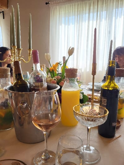 The scene depicts an indoor dining or event setting in Favões, Portugal, focusing on a long table covered with a white tablecloth. In the foreground, a stemmed glass containing rosé wine stands prominently next to a partially visible clear glass with water. To its right, a crystal coupe glass with a gold rim holds small, light-colored items.

In the midground, a silver ice bucket contains at least one dark-colored wine bottle. Nearby, various clear glass bottles are present: one with orange juice, others with cork stoppers containing liquids with fruit slices and herbs, and one with a pink cap holding flowers. A dark-colored wine bottle with a label, possibly red wine, rests directly on the tablecloth. Floral arrangements featuring white tulips, yellow flowers, and pink roses are interspersed among the bottles. Decorative elements include a brass candelabra holding four unlit green taper candles and a single brass candle holder with an unlit red taper candle.

Two individuals are partially visible at the table. On the left, a woman with dark hair is seen in profile, looking downwards or to the side. On the right, another woman with dark, shoulder-length hair is partially visible, appearing to look towards the table. The background features a large window draped with sheer white curtains, providing diffused lighting, and a light-colored wall.