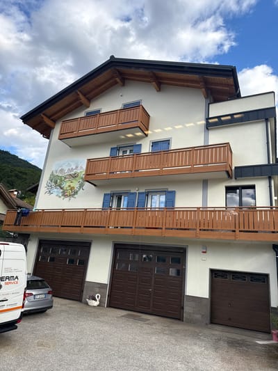 This media file captures an exterior view of a multi-story building, likely a residential or commercial property, in Cavedago, Italy. The building features a light-colored facade with several brown wooden balconies and shutters. A prominent artistic mural is visible on the facade, depicting a scenic landscape with mountains, a village, and a body of water.
The scene is set outdoors, with a cloudy sky overhead suggesting overcast weather. The ground is paved, and two dark brown garage doors are visible on the lower level of the building. A silver car with the license plate "EY-246VK" is parked in front of one of the garages, alongside a white van. The van has text on its side, including "MODERMA.IT", "MODERMA SRL-TRENTO", "ERMA SERVICE", and "RICAMBI NOLEGGIO". A white swan-like decoration is placed near the garages. The overall impression is that of a residential area in a mountainous region, possibly during daytime.