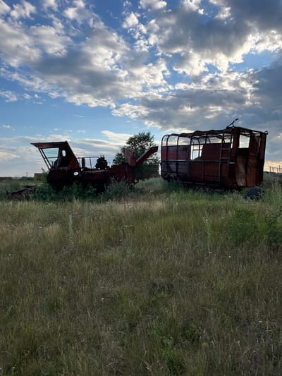 In the city of Bayagiz, Kazakhstan, an abandoned farmstead is depicted. The scene is set in a grassy field under a cloudy sky. In the foreground, tall, dry grass with scattered small wildflowers covers the ground. Two rusted agricultural machines stand in the mid-ground. The machine on the left appears to be a harvester with a visible cab and mechanical components, partially overgrown with green foliage. To its right is a large, open-topped trailer or wagon, also heavily rusted and with a skeletal frame. The background shows more distant vegetation and a treeline. The overall impression is one of decay and abandonment in a rural setting.