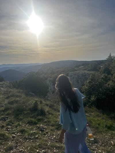 A woman with long dark hair stands on rocky, grassy terrain, possibly a cliffside or elevated area, near Bonnieux, France. She is wearing a light blue top and blue pants. In her hand, she is holding a plastic bottle containing a yellow liquid. The background shows a mountainous landscape under a bright sky, possibly during sunrise or sunset due to the angle and intensity of the sunlight. The environment consists of green vegetation and rocky surfaces.