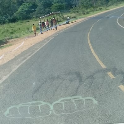The image displays a roadside scene in Kola ward, Kenya, captured from within a vehicle. An asphalt road, featuring a solid yellow center line and a broken white shoulder line, curves to the right and recedes into the distance. A dirt/gravel shoulder separates the road from a grassy bank on the left. In the background, a hillside is densely covered with green trees and vegetation.

Approximately eight individuals are present on the dirt shoulder, primarily gathered near a rudimentary, open-sided shelter with a light-colored roof. One individual, wearing a yellow shirt and dark trousers, is walking away from the main group towards the background on the shoulder. The remaining seven individuals are standing under or adjacent to the shelter. A light-colored motorcycle or scooter rests on its side near the shelter, partially obscured by the standing people. Abstract light-colored patterns, consistent with reflections or markings on a vehicle windshield, are visible in the lower foreground of the image. Darker, indistinct patterns are also present on the road surface, likely shadows or reflections.