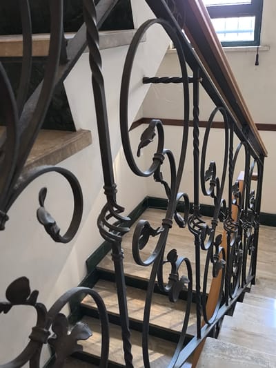 The image shows a close-up view of a wrought iron staircase railing with ornate scrollwork and twisted balusters. The railing is dark gray or black and appears to be made of metal. The stairs themselves are made of marble with light and dark veining. The wall behind the railing is a light color, possibly white or off-white. A wooden handrail runs along the top of the railing. A window with multiple panes is visible at the top right of the image, with a dark green frame. The scene is indoors, likely in a building with traditional architecture. There are no people or animals visible in the image.