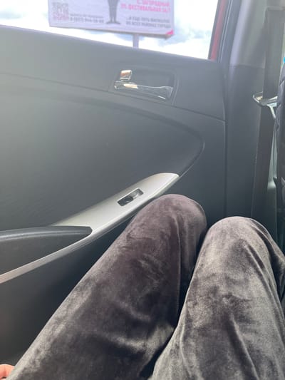 The image shows the interior of a car. In the foreground, a person's legs, clad in dark gray velour pants, are visible. The pants are creased, indicating the person is sitting. The car's interior includes a black door panel with a silver trim and a chrome door handle. A silver-colored window control panel is visible on the door. Beyond the car's interior, a blurry billboard with text in Russian is visible through the car window, suggesting an outdoor urban or suburban environment. The billboard appears to have a QR code and phone numbers. The lighting suggests it is daytime. The location is identified as Novaya Otradovka, Russia.