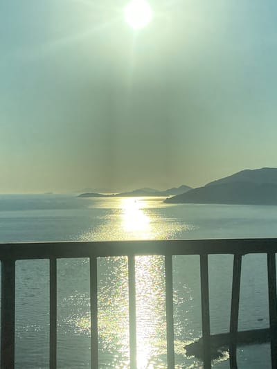 This image captures a serene view of the Adriatic Sea from Plat, Croatia. The sun is high in the sky, casting a bright, shimmering reflection across the water, which is calm with gentle ripples. In the distance, hazy islands and mountains form a silhouetted landscape against the soft, light blue sky. A dark, metallic railing in the foreground frames the scene, with its vertical bars creating a pattern that partially obscures the view of the water. The overall impression is one of tranquil beauty, with the sunlight and the vast expanse of the sea dominating the composition.