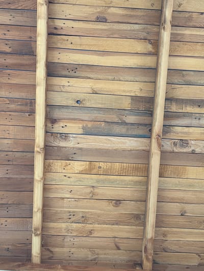 The image shows a close-up, overhead view of a wooden ceiling constructed from horizontal planks. Two vertical wooden beams are present, running from the top to the bottom of the frame. The planks exhibit variations in color, ranging from light tan to dark brown, and show visible wood grain, knots, and nail holes. The overall texture is rough and natural. The scene is located outdoors, likely under a covered structure, given the natural lighting and the unfinished appearance of the wood. There are no people or any other objects visible in the image. The location is identified as Golyam Chardak, Bulgaria.