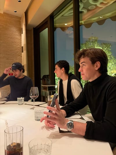 Three young men are seated at a table in a restaurant. One man, wearing a black shirt, is using a cell phone with a stainless steel wristwatch visible on his wrist. Another man, wearing a white shirt with a black jacket, is looking down, presumably at his own device. A third man, wearing a dark baseball cap and a dark shirt, appears to be on a phone call. The table is set with various glasses and what may be menus. The restaurant has a modern interior with a window view.