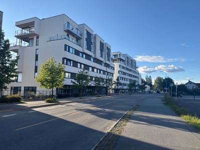 The image captures a street scene in Jessheim, Norway, likely on a bright, sunny day with a clear blue sky and scattered clouds. In the foreground, an asphalt road with yellow and white lane markings is visible, casting long shadows from the surrounding buildings and trees. To the left, a modern, multi-story apartment building with a white facade, dark-trimmed windows, and glass balconies stretches along the street. Trees and small bushes are planted along the sidewalk in front of the building.

The apartment building appears to be part of a development, with several interconnected sections visible, each featuring a similar architectural style with protruding sections and balconies. Further down the street, more of the apartment buildings are visible, receding into the distance. A few cars are present on the road, and a pedestrian is seen walking on the sidewalk.

To the right of the road, there is a sidewalk and a grassy verge. In the background, more trees and some residential houses are visible, suggesting a suburban or semi-urban setting. The overall impression is one of a contemporary urban or semi-urban residential area.