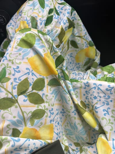 The image shows a close-up view of a white fabric with a repeating pattern of lemons, green leaves, and blue floral motifs. The fabric is gathered and draped, suggesting it might be part of a garment or home decor item. The background is dark and out of focus. The fabric is likely located within Skra, Georgia, although no specific landmarks are visible in the image to confirm this location. There are no people or actions depicted.