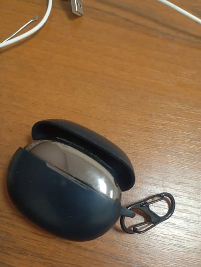The image shows a close-up, angled view of a dark blue silicone case for wireless earbuds, lying on a wooden surface. The case is open, revealing the metallic, silver-colored earbuds inside. Attached to the bottom of the case is a black carabiner clip, designed for easy attachment to bags or belts. The wooden surface has a visible grain pattern, suggesting a desk or table. In the upper left corner of the image, a portion of a white cable and a silver USB connector are visible, hinting at charging equipment. The lighting suggests an indoor setting, possibly during the day, with no specific weather cues. There is no visible text in the image. The scene is static, with no discernible activity or event taking place; it appears to be a product shot or a personal item resting on a surface.
