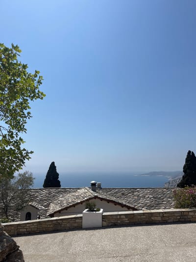 The image shows a view of the Aegean Sea from the grounds of the Monastery of Archangel Michael in Moni Archaggelou, Greece. The image was taken from the coordinates 40.597, 24.701. The monastery is located on a hilltop with a stone wall surrounding the grounds. In the foreground, there is a stone patio with a white planter holding a bush. The view from the patio shows the Aegean Sea with the coastline in the distance. There are several small trees and shrubs in the foreground, as well as the roof of the monastery building, with one chimney visible. The sky is clear and blue.