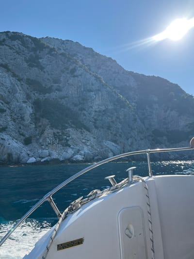 The image shows a view from a boat moving on the Aegean Sea towards the mountainous coastline of Skopelos, Greece. The water is a deep blue with white foam generated by the boat's movement. The sun shines brightly from the upper right of the frame, casting a glare on the water and partially obscuring the details of the rocky, vegetated mountainside. The boat's white hull and a metal railing are visible in the foreground. A coiled rope is secured to a cleat on the deck. A yellow warning label with text in Greek and English, indicating a slippery surface when wet, is attached to the side of the boat. The overall impression is one of a sunny day at sea with a dramatic coastal landscape.