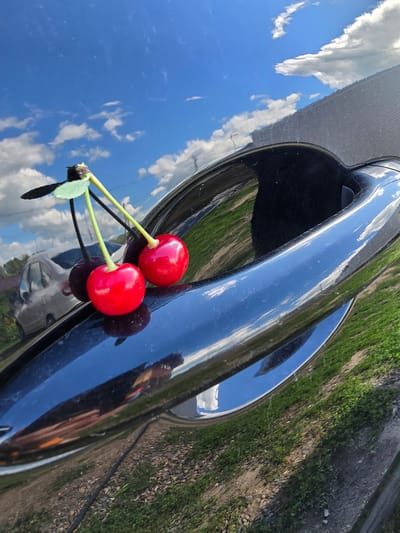Two artificial cherries are placed on the black, reflective surface of a car door handle. The cherries are red with green stems, and a small green leaf is attached to one of the stems. The reflection in the car shows a bright blue sky with white clouds, green grass, and a silver car. The image was taken in Ермолаевский Затон, Russia.