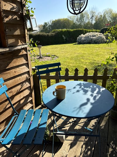 Outdoor scene in Hitchin, United Kingdom. A blue metal bistro set with a round table and two chairs sits on a wooden deck. A yellow mug rests on the table. A wooden fence separates the deck from a green lawn with various trees and bushes. On the left is a wooden structure with a solar light on top. The lighting suggests it is daytime.
