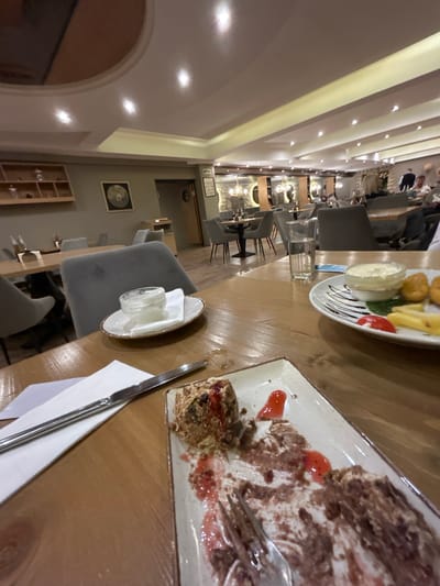 The image captures a long table in a restaurant, likely in Pamporovo, Bulgaria. The table is laden with remnants of a meal, including a plate with a partially eaten dessert, possibly a chocolate cake with cream and fruit sauce. Another plate contains fried items, such as fries and nuggets, accompanied by dipping sauce and tomato slices. A glass of water and a small dish with what appears to be a condiment or sauce sit nearby. Cutlery and a napkin are also present.
The restaurant is filled with chairs and tables. In the background, indistinct figures of people can be seen. The interior design includes archways, a shelf with glassware, and a framed circular design on the wall.