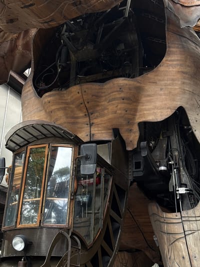 The image captures a close-up view of a section of the Great Elephant of Nantes, a mechanical sculpture located in the Machines de l'île on the Île de Nantes in Nantes, France. The sculpture is constructed primarily of wood and metal. The foreground features the intricately designed cabin of the elephant, with its large glass windows and wooden framing. A prominent headlight is visible on the lower left. Behind the cabin, the wooden exterior of the elephant's body is seen, with its characteristic undulating shape and visible rivets. Openings in the wooden structure reveal the complex mechanical workings within, including dark metal components, pipes, and wires. The overall impression is one of a massive, detailed, and somewhat industrial work of art, set against what appears to be an outdoor or covered exhibition space.