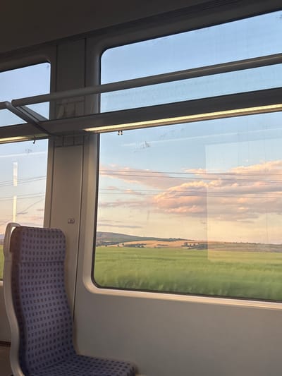 The image depicts the interior of a train carriage, specifically focusing on a window view of a rural landscape during the late afternoon or early evening, consistent with the golden hour. The scene is located in Tsar Asen, Bulgaria.
In the foreground, an empty train seat occupies the lower left quadrant. The seat is upholstered in a blue fabric featuring a repeating dark blue square grid pattern, with a light grey plastic frame. A narrow vertical panel separates the front window from a larger main window; this panel has a small, rectangular metallic sign affixed to it, displaying the number "55" in black. Overhead, the train's interior features grey panels and horizontal lighting strips that emit a warm, yellow light. The window glass exhibits some dirt, smudges, and faint reflections of the train's interior structure and power lines from outside.
Through the main window, an expansive outdoor landscape is visible. The sky transitions from a light blue at the top to warmer hues of orange and pink closer to the horizon, indicative of sunset or sunrise. Scattered, puffy clouds are present, reflecting the warm light. Across the middle ground, multiple dark, horizontal power lines stretch across the frame. The terrain beyond consists of rolling hills and vast agricultural fields, some appearing bright green and others displaying drier, golden-brown tones. Clusters of trees are interspersed throughout the fields and on the distant hillsides. There are no individuals present or visible actions occurring within the frame.