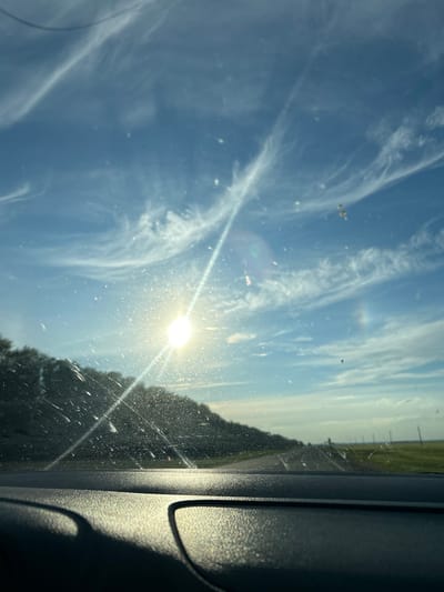 The image shows a bright, sunlit sky with wispy clouds, viewed through what appears to be a dusty or streaked window, possibly from inside a vehicle. The sun is a prominent, almost blindingly bright orb, casting a strong lens flare across the scene and illuminating small particles in the air. In the distance, a flat landscape with a field and possibly some trees or vegetation can be seen under a pale sky. The right side of the image is dominated by the dark, textured interior of a vehicle, likely a car's dashboard or pillar, with harsh shadows and highlights. The overall impression is one of a sunny day, possibly during travel, with the sun's glare creating a somewhat abstract and bright atmosphere. There is no visible text. The orientation of the image suggests it was taken in portrait mode and possibly rotated. Given the location context of Ilteryakovo, Russia, it depicts a scene typical of a rural or roadside view on a clear day.